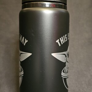 May include: Black water bottle with a black lid and handle. The bottle features a white graphic of a character from a popular science fiction series, along with the text "THIS IS THE WAY".