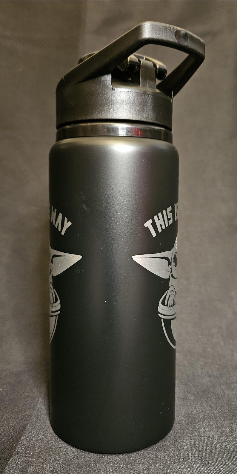 May include: Black water bottle with a black lid and handle. The bottle features a white graphic of a character from a popular science fiction series, along with the text "THIS IS THE WAY".
