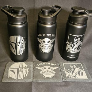 May include: Three black water bottles with silver designs and matching coasters. The bottles feature images of a Mandalorian helmet, Baby Yoda, and a Mandalorian character. The coasters have the same designs.