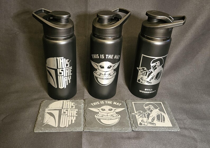 May include: Three black water bottles with silver designs and matching coasters. The bottles feature images of a Mandalorian helmet, Baby Yoda, and a Mandalorian character. The coasters have the same designs.