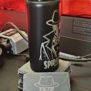 Peut inclure: Une bouteille d'eau en acier inoxydable noir mat avec un couvercle noir et une gravure laser repr&eacute;sentant un personnage en trench-coat et chapeau fedora avec le texte SPIDER NOIR, accompagn&eacute;e d'un dessous de verre carr&eacute; en ardoise assorti.