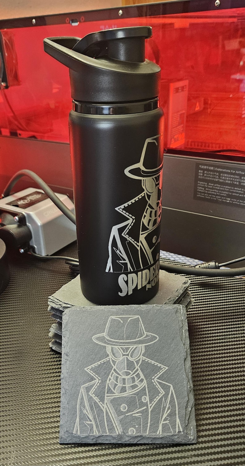 Peut inclure: Une bouteille d'eau en acier inoxydable noir mat avec un couvercle noir et une gravure laser repr&eacute;sentant un personnage en trench-coat et chapeau fedora avec le texte SPIDER NOIR, accompagn&eacute;e d'un dessous de verre carr&eacute; en ardoise assorti.