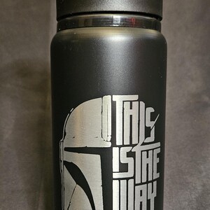 May include: Black water bottle with a black lid. The bottle features a silver graphic of a Mandalorian helmet and the phrase "THIS IS THE WAY". The bottle is designed for cold beverages.