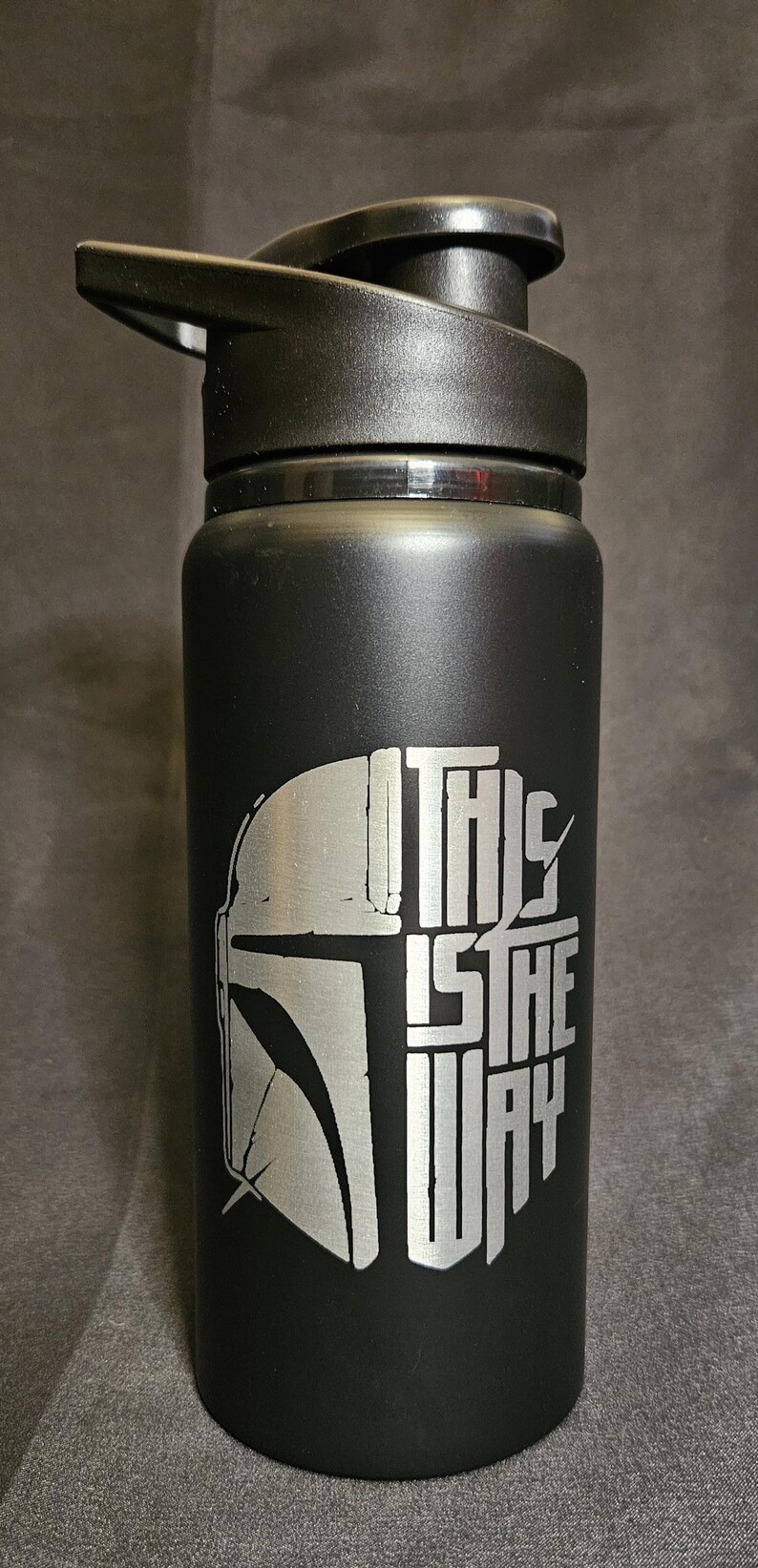 May include: Black water bottle with a black lid. The bottle features a silver graphic of a Mandalorian helmet and the phrase "THIS IS THE WAY". The bottle is designed for cold beverages.