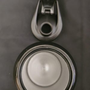 May include: An overhead shot of a stainless steel tumbler with a black rim and a matching lid. The lid is open, revealing the interior of the tumbler. The background is a solid gray.