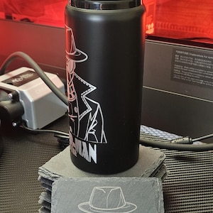 Peut inclure: Une bouteille d'eau noire mate avec un couvercle noir et un graphisme au trait blanc repr&eacute;sentant un personnage portant un fedora et un trench-coat, avec le texte SPIDER-MAN en dessous. Un dessous de verre en ardoise assorti porte le m&ecirc;me motif.