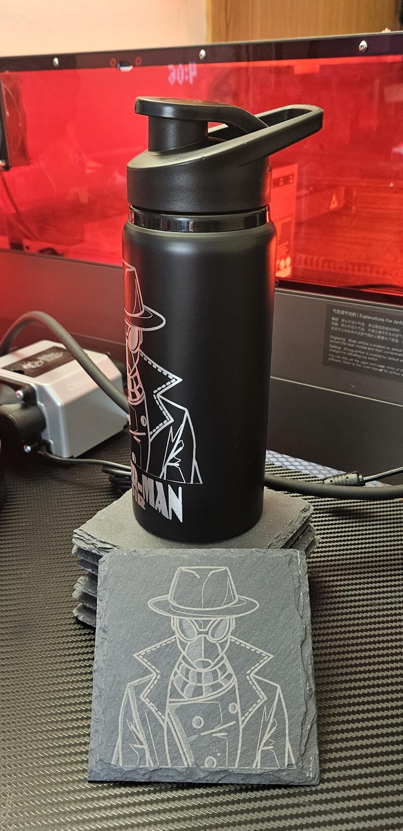 Peut inclure: Une bouteille d'eau noire mate avec un couvercle noir et un graphisme au trait blanc repr&eacute;sentant un personnage portant un fedora et un trench-coat, avec le texte SPIDER-MAN en dessous. Un dessous de verre en ardoise assorti porte le m&ecirc;me motif.