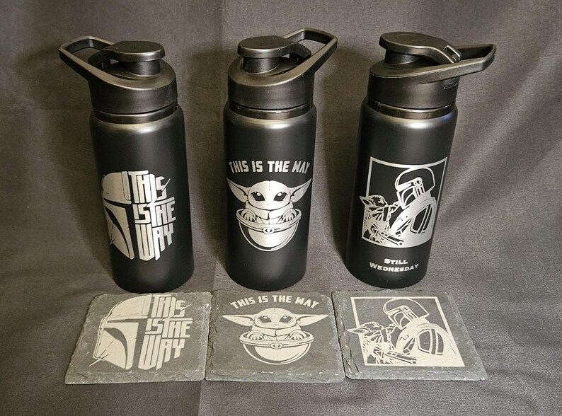May include: Three black water bottles with silver designs and matching coasters. The bottles feature images of a helmet, a character, and the text "THIS IS THE WAY". The coasters have the same designs. The bottles are approximately 9 inches tall.