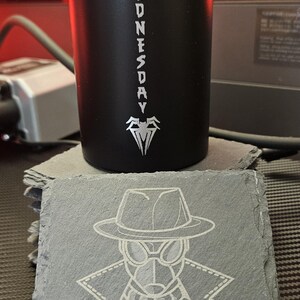 Peut inclure: Un gobelet cylindrique noir mat avec le texte vertical blanc STILL WEDNESDAY et un embl&egrave;me d'araign&eacute;e. Devant se trouve un dessous de verre carr&eacute; en ardoise grav&eacute; d'une illustration au trait blanc d'un personnage portant un fedora et un trench.
