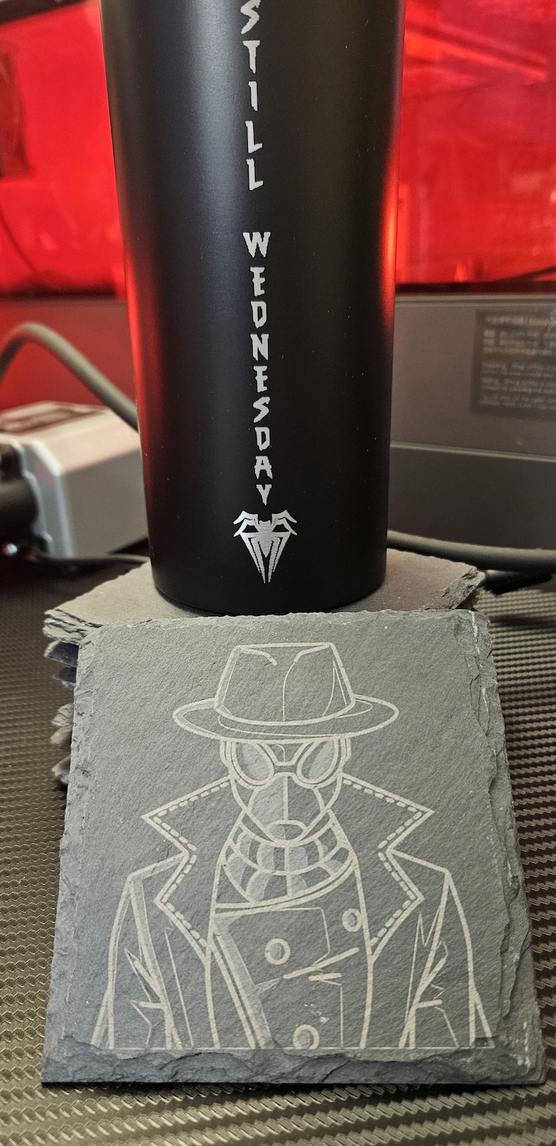 Peut inclure: Un gobelet cylindrique noir mat avec le texte vertical blanc STILL WEDNESDAY et un embl&egrave;me d'araign&eacute;e. Devant se trouve un dessous de verre carr&eacute; en ardoise grav&eacute; d'une illustration au trait blanc d'un personnage portant un fedora et un trench.