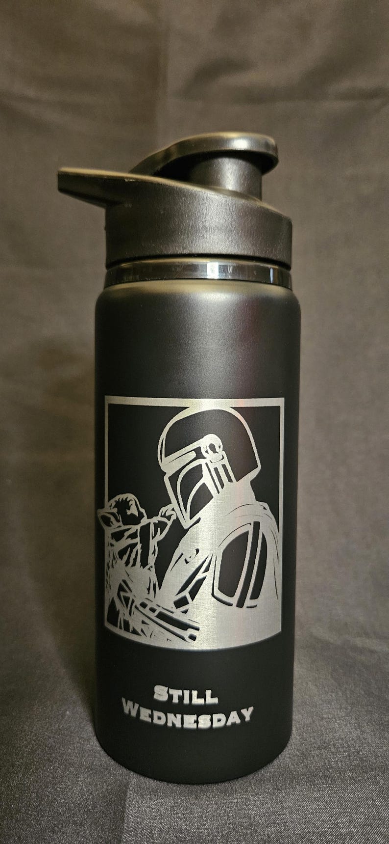 May include: Black water bottle with a flip-top lid. The bottle features a silver graphic of a Mandalorian and Grogu, with the text "STILL WEDNESDAY" below the graphic. The bottle is designed for cold beverages.