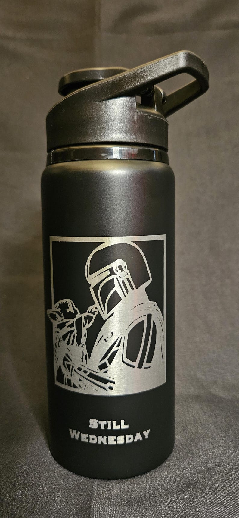 May include: Black water bottle with a flip-top lid. The bottle features a silver graphic of a character from a popular science fiction series, holding a small creature. The text "STILL WEDNESDAY" is below the graphic.
