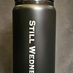 May include: Black water bottle with a black flip-top lid. The words "STILL WEDNESDAY" are printed vertically in white on the side of the bottle. The bottle is cylindrical and appears to be made of stainless steel.