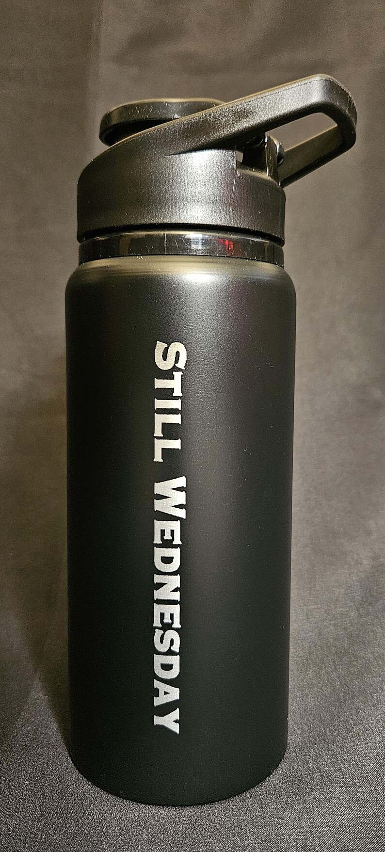 May include: Black water bottle with a black flip-top lid. The words "STILL WEDNESDAY" are printed vertically in white on the side of the bottle. The bottle is cylindrical and appears to be made of stainless steel.