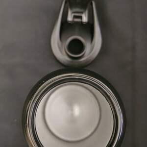 Peut inclure: Une vue de dessus d'une bouteille d'eau noire ouverte avec un int&eacute;rieur en m&eacute;tal argent&eacute;. Le couvercle en plastique noir d&eacute;tach&eacute; pr&eacute;sente une ouverture circulaire et un m&eacute;canisme de bouchon &agrave; charni&egrave;re, plac&eacute; au-dessus de l'ouverture.