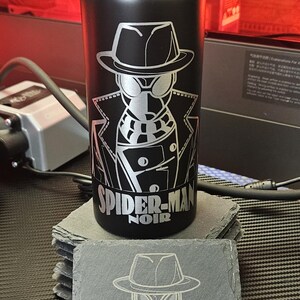 Peut inclure: Une bouteille d'eau noire mate avec un bouchon noir et une gravure laser argent&eacute;e repr&eacute;sentant Spider-Man Noir. Sous le motif, le texte indique SPIDER-MAN NOIR. Un dessous de verre en ardoise carr&eacute;e assorti avec le m&ecirc;me motif est pos&eacute; devant.