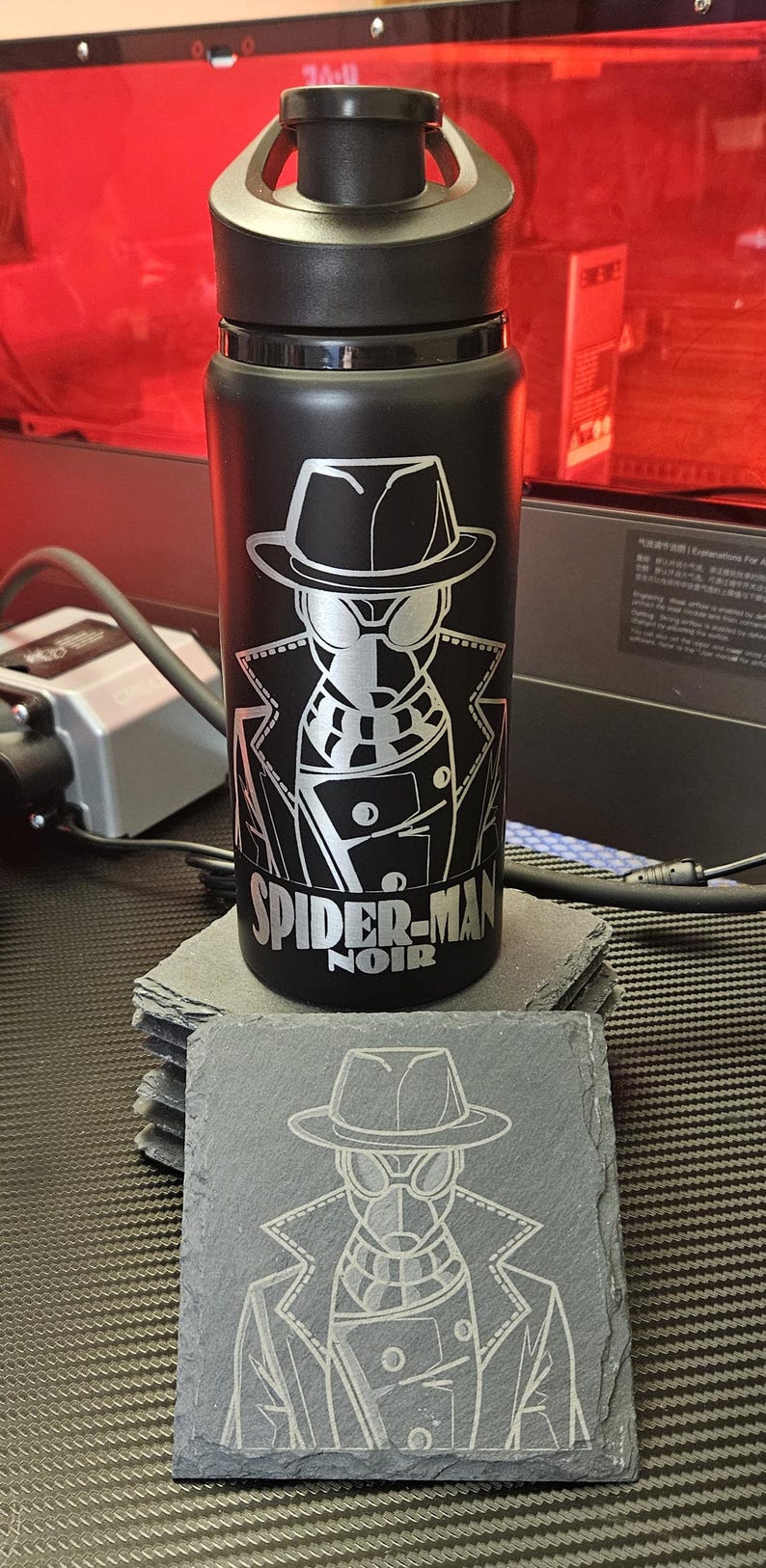Peut inclure: Une bouteille d'eau noire mate avec un bouchon noir et une gravure laser argent&eacute;e repr&eacute;sentant Spider-Man Noir. Sous le motif, le texte indique SPIDER-MAN NOIR. Un dessous de verre en ardoise carr&eacute;e assorti avec le m&ecirc;me motif est pos&eacute; devant.