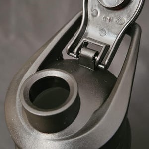 May include: Close-up of a black water bottle with a flip-top lid. The lid is open, revealing the drinking spout. The bottle is made of a matte black material and has a sleek, modern design. The bottle is designed for sports and outdoor activities.