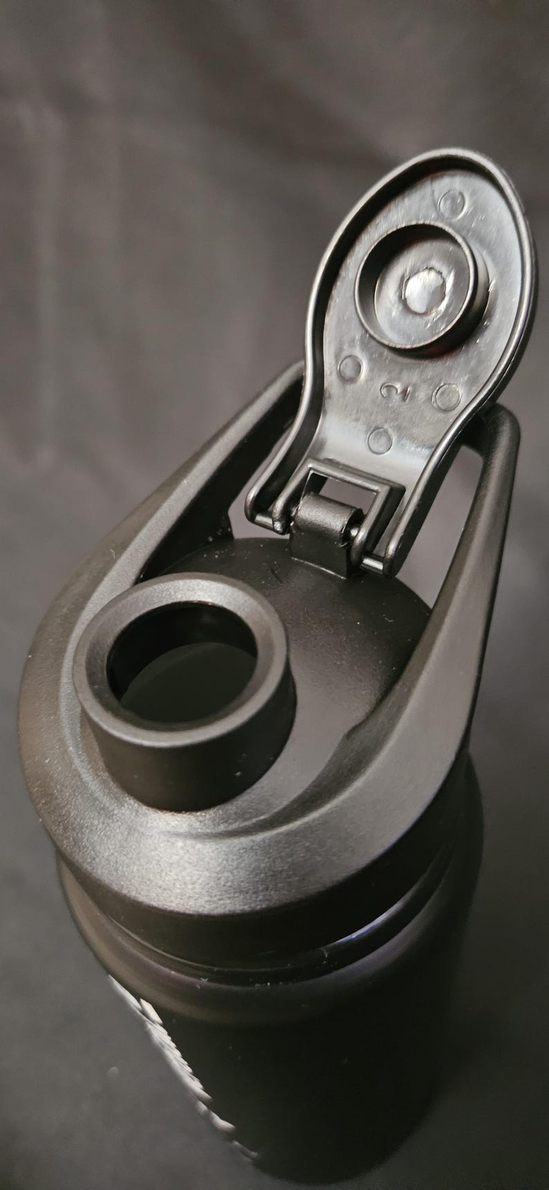 May include: Close-up of a black water bottle with a flip-top lid. The lid is open, revealing the drinking spout. The bottle is made of a matte black material and has a sleek, modern design. The bottle is designed for sports and outdoor activities.