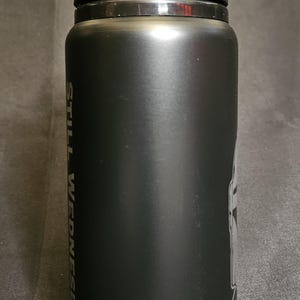 May include: Black insulated water bottle with a black handle and lid. The bottle has the words "STILL WEDNESDAY" printed vertically in white. A graphic is visible on the side.