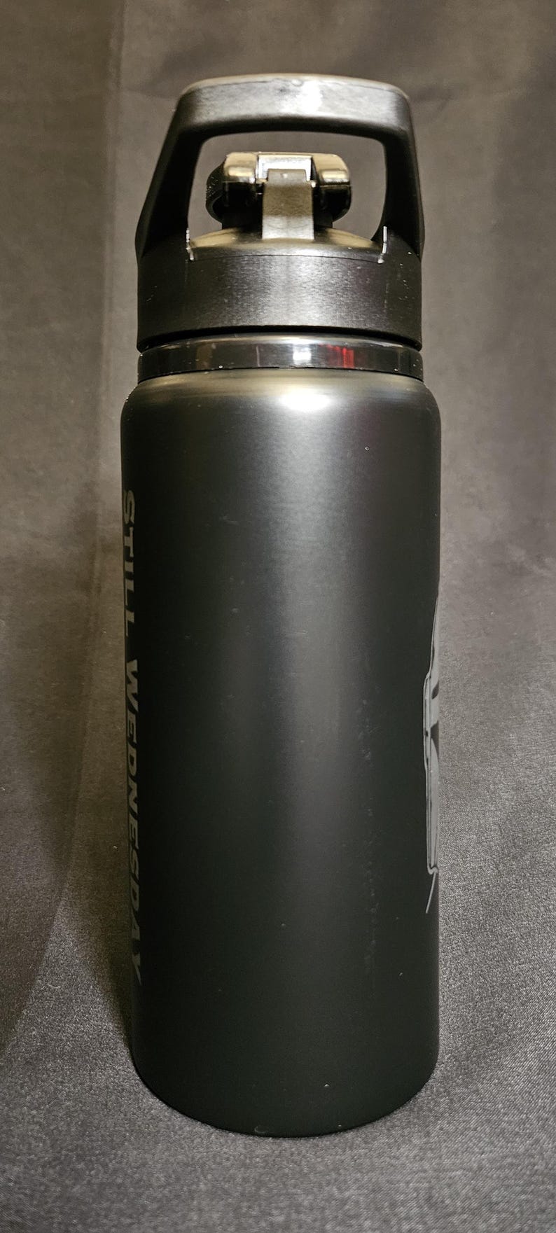 May include: Black insulated water bottle with a black handle and lid. The bottle has the words "STILL WEDNESDAY" printed vertically in white. A graphic is visible on the side.