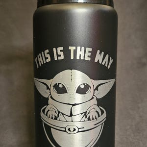 May include: Black water bottle with a silver engraving of a character from a popular science fiction franchise. The text "THIS IS THE WAY" is above the character. The bottle has a black handle.