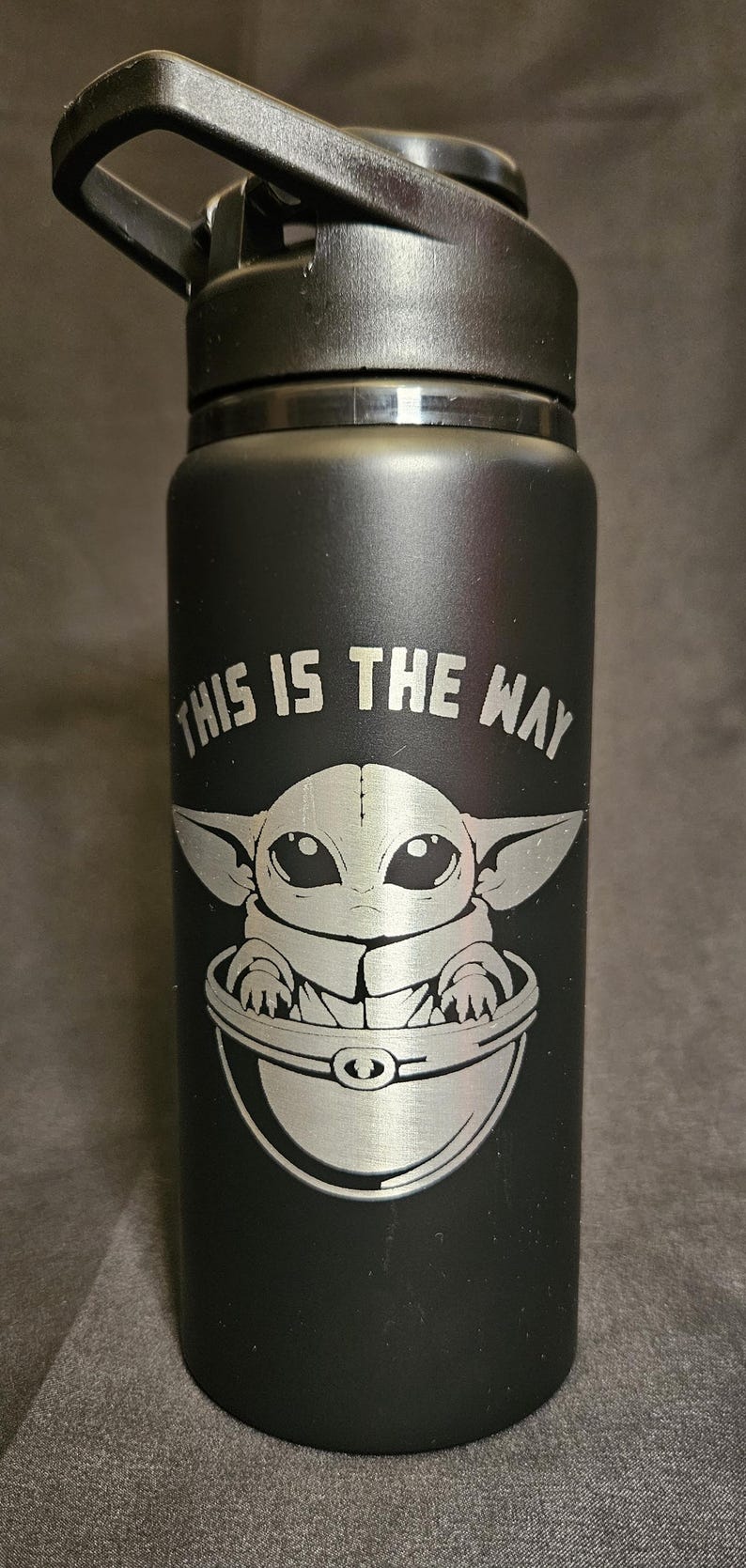 May include: Black water bottle with a silver engraving of a character from a popular science fiction franchise. The text "THIS IS THE WAY" is above the character. The bottle has a black handle.