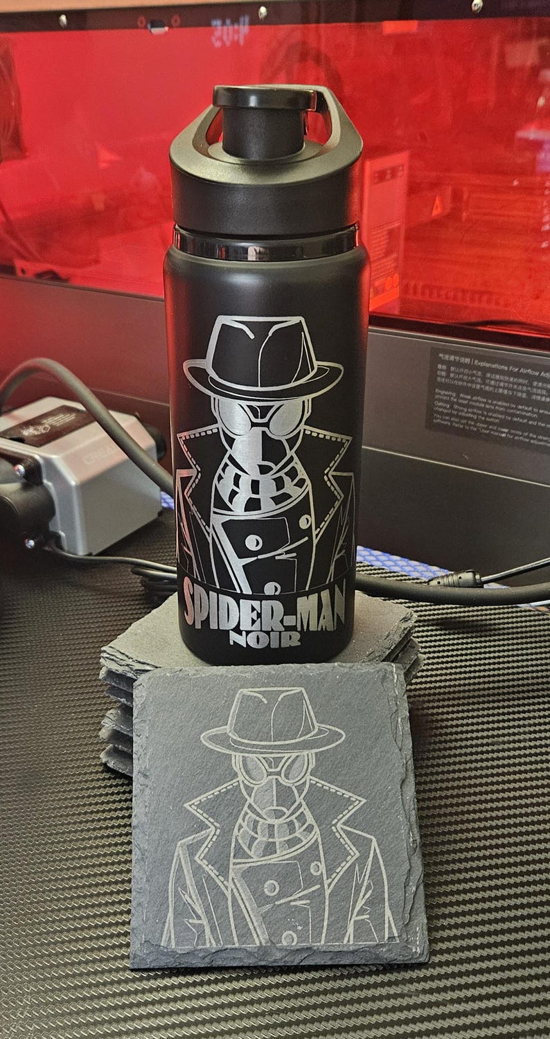 Peut inclure: Une bouteille d'eau en m&eacute;tal noir avec un bouchon &agrave; vis noir, pr&eacute;sentant une gravure laser du personnage Spider-Man Noir et le texte "SPIDER-MAN NOIR" en blanc, accompagn&eacute;e d'un dessous de verre carr&eacute; en ardoise avec le m&ecirc;me motif grav&eacute;.