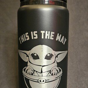 May include: Black water bottle with a black lid. The bottle features a silver engraving of Grogu, also known as Baby Yoda, in a pod, with the phrase "THIS IS THE WAY" above the image.