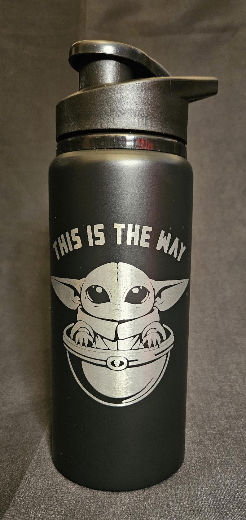 May include: Black water bottle with a black lid. The bottle features a silver engraving of Grogu, also known as Baby Yoda, in a pod, with the phrase "THIS IS THE WAY" above the image.