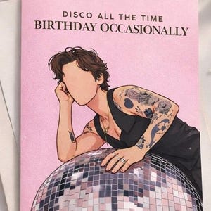 May include: Pink birthday card with the text "DISCO ALL THE TIME BIRTHDAY OCCASIONALLY". The card features an illustration of a person with tattoos, resting on a disco ball. The card is on a white surface.