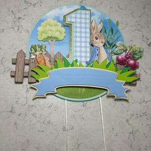 May include: A Peter Rabbit themed cake topper featuring a blue and white gingham number one, a rabbit in a blue jacket, and a blue banner. The topper includes a wooden fence, a tree, and vegetables.