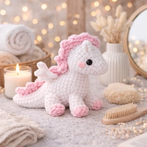May include: A handmade crochet unicorn toy in white and pink. The unicorn has a pink mane, tail, and hooves, with small white wings. It features black eyes and pink cheeks. The toy is sitting on a light grey surface.