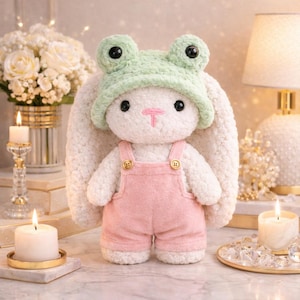 Op de afbeelding: Een pluche wit konijn met een lichtgroene kikkerhoed en een roze overall. Het konijn heeft zwarte ogen en een roze neus. De overall heeft goudkleurige knopen. Het konijn staat op een marmeren oppervlak.