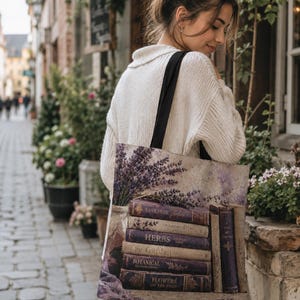Könnte beinhalten: Eine Tragetasche mit schwarzen Trägern und einem Aufdruck, der einen Lavendelstrauß in einer Vase neben einem Stapel alter Bücher mit den Titeln Lavender Fields, Herbs, The Garden, Botanical Illustrations und Flowers of the Field zeigt.