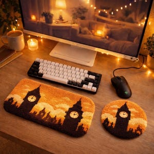 Puede incluir: Una configuración de escritorio con un teclado en blanco y negro, un ratón negro y un tapete y posavasos con el tema del Big Ben. El tapete y el posavasos tienen un diseño de puesta de sol en marrón y naranja. También se ve un monitor de ordenador y una taza de café.
