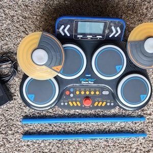 May include: A black VTech KidiStar Drum Pad featuring four circular grey pads, two translucent yellow cymbals, a central control panel with an orange volume knob, a digital display, two blue drumsticks, and a black power adapter with a cord.