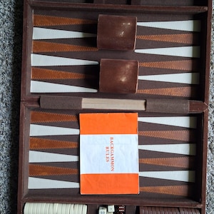 Puede incluir: Juego de backgammon abierto con una superficie de juego marrón, beige y blanca. El juego incluye cubiletes marrones, dados y fichas. Se ve un folleto naranja con el texto "BACKGAMMON RULES".