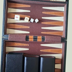 May include: A black backgammon set featuring a brown felt interior with alternating cream and brown triangular points. The set includes brown and cream checkers, two black dice cups, two standard dice, and a doubling cube marked with the number 32.