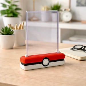 Op de afbeelding: Een rode, witte en zwarte bureaulamp met een helder rechthoekig lichtpaneel. De lamp heeft een ronde knop en is ontworpen om op een Pokeball te lijken. De lamp staat op een houten bureau.