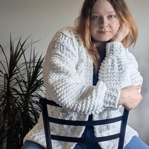 May include: A chunky, white, long-sleeved cardigan made from a thick, soft, velvet-like yarn. The garment features a textured, cable-knit pattern with ribbed cuffs and an open front design, providing a cosy, layered look over a dark shirt and blue jeans.