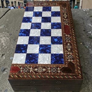 May include: A wooden chess set featuring a blue and white chequered playing surface. The board is framed with detailed inlaid wood designs in brown and red hues. The closed box is ready for play or travel.