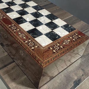 Könnte beinhalten: Eine Holzschachkiste mit Schachbrettmuster. Die Oberseite ist schwarz und weiß, mit einem dekorativen Rand aus eingelegtem Holz und geometrischen Mustern. Die Box ist aus dunklem Holz mit eingelegten Designs gefertigt.