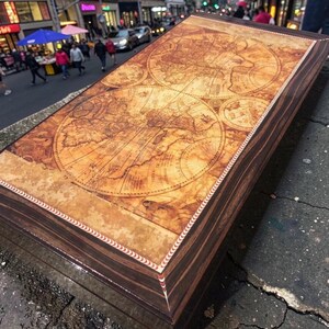 Könnte beinhalten: Eine rechteckige Holzschatulle mit einer auf den Deckel gedruckten Weltkarte im Vintage-Stil. Die Schatulle hat Seiten aus dunkelbraunem Holzmaserungsmuster und einen dekorativen Rand mit einem rot-weißen Muster um die zentrale Kartenabbildung.