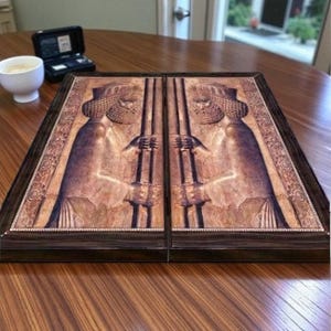 May include: A rectangular wooden backgammon board featuring a dark brown frame and a central inlay depicting two mirrored, ancient-style figures with detailed beards and headwear. The board surface displays a warm, textured, tan-colored relief design.