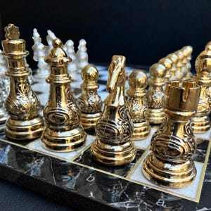 Könnte beinhalten: Ein Schachspiel mit polierten, goldfarbenen Metallfiguren, die mit aufwendigen, geprägten Schnörkelmustern verziert sind. Das Set enthält König, Dame, Springer und Turm auf einem schwarz-weiß marmorierten Brett mit sichtbaren Buchstaben.