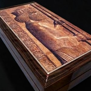 May include: A rectangular wooden box featuring a detailed relief carving on its lid. The carving depicts a figure in profile wearing a textured headdress and holding a long staff. The border is decorated with a repeating floral pattern.