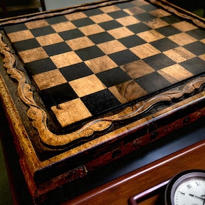 Peut inclure: Un échiquier carré en bois présentant une grille de carrés alternés brun clair et noir. Le plateau est entouré d'un cadre en bois épais et sculpté avec un motif de bordure ondulé et décoratif, mettant en valeur les textures du grain.