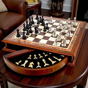 Peut inclure: Un jeu d'échecs en bois avec un plateau carré composé de cases claires et foncées, bordé d'une incrustation décorative. Le jeu comprend des pièces en métal noir et argenté sur le plateau et un tiroir coulissant contenant des pièces dorées.