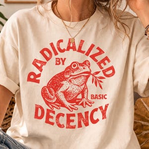 Puede incluir: Camiseta color crema con un gráfico rojo de una rana sosteniendo una ramita. El texto "RADICALIZED BY BASIC DECENCY" está impreso en un diseño circular alrededor de la rana. La camiseta tiene mangas cortas y un corte holgado.